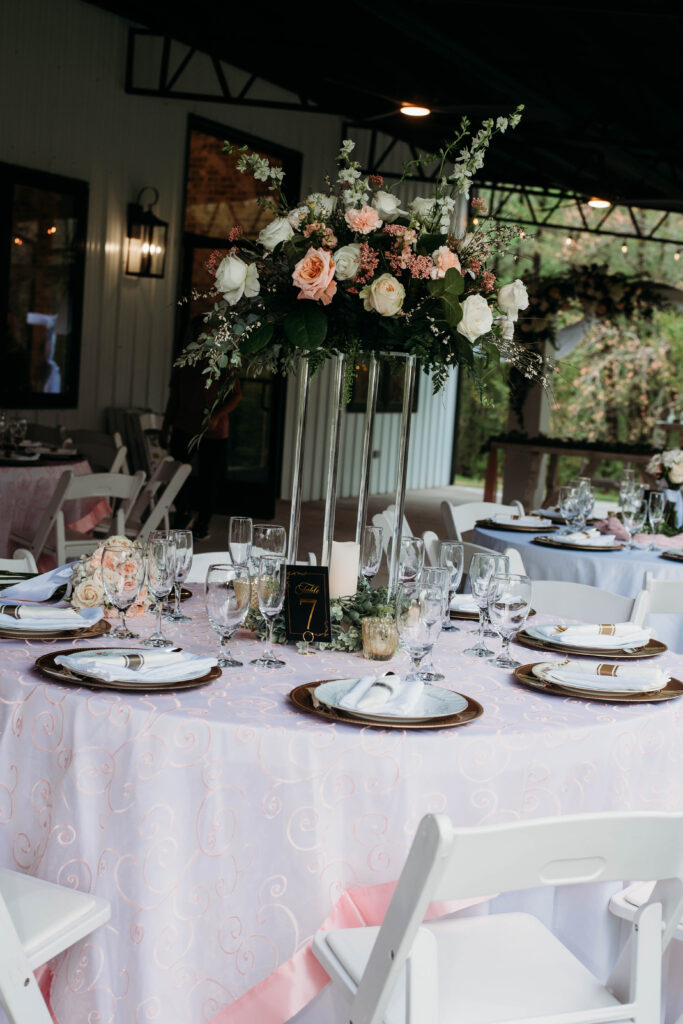 A sophisticated outdoor reception setup featuring Table 2. The round table is adorned with a delicate pink patterned tablecloth, gold-rimmed chargers, and crisp white napkins with gold accents. A tall, modern clear stand serves as the centerpiece, showcasing a lush, romantic floral arrangement of blush roses and soft greenery. Crystal glassware and wine bottles complete the formal place settings, while the covered patio venue offers a beautiful, natural wooded backdrop for an upscale wedding atmosphere.