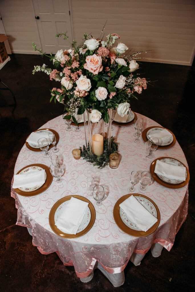 An overhead view of a circular wedding reception table fully set for six guests. The table features a pale pink, swirling patterned overlay, gold-rimmed chargers, and delicate floral china. The centerpiece is a tall, clear acrylic stand topped with a lush, romantic arrangement of blush pink and ivory roses, surrounded by greenery and soft candlelight. This setup showcases our elegant event styling services, highlighting our attention to detail in creating sophisticated, classic wedding tablescapes for our clients.