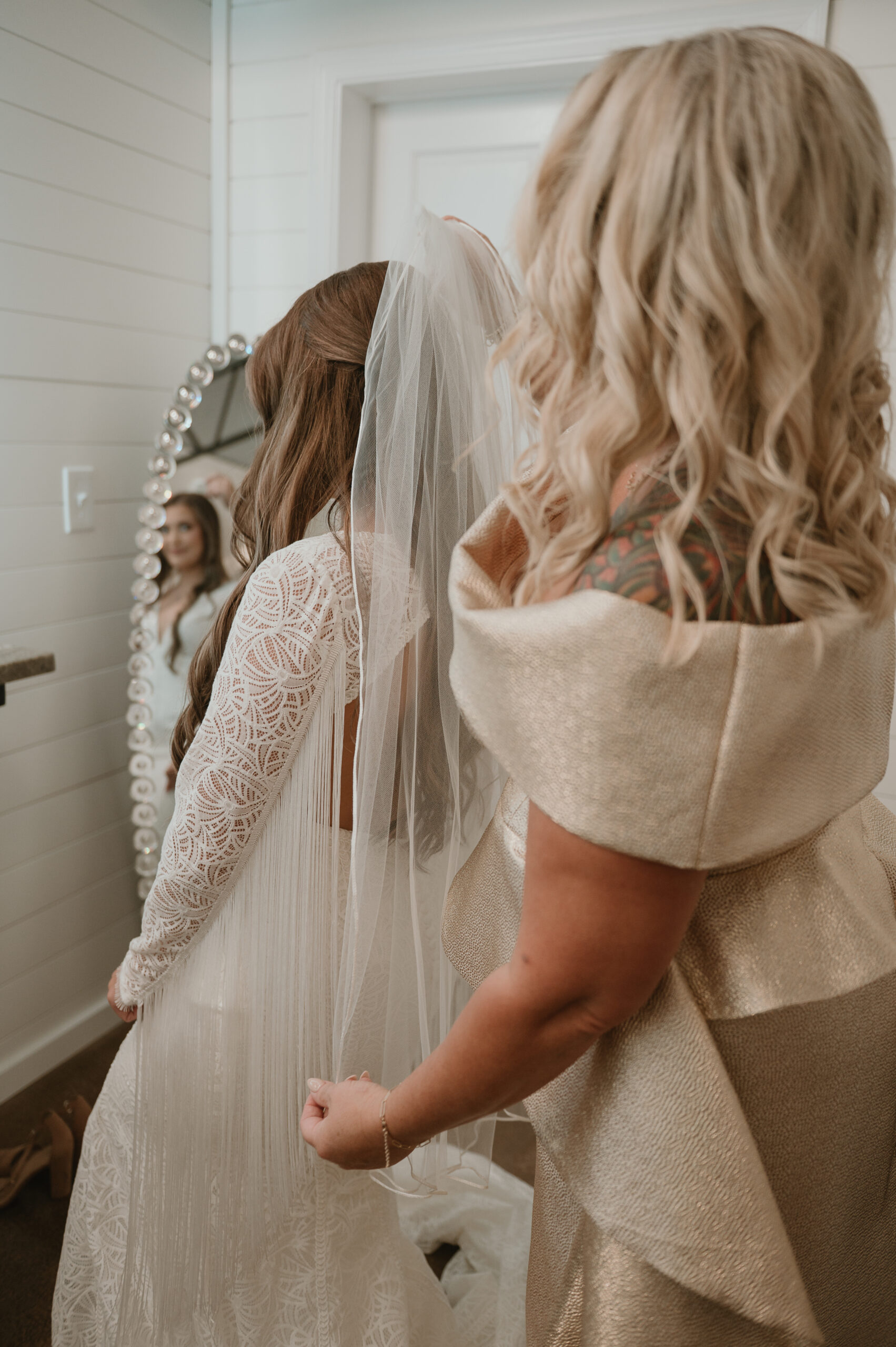 A behind-the-scenes shot of a bride wearing a lace fringe gown while a loved one adjusts her veil, captured in a boutique vanity mirror reflection.