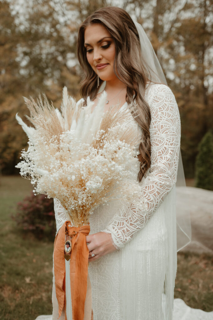 A bride in a bohemian lace gown holding a custom-designed, textural dried bouquet. The arrangement features fluffy pampas grass and delicate baby’s breath, tied with a burnt orange ribbon and a sentimental memory locket. This shot highlights the soft, earthy color palette and preserved floral elements we offer for fall weddings.