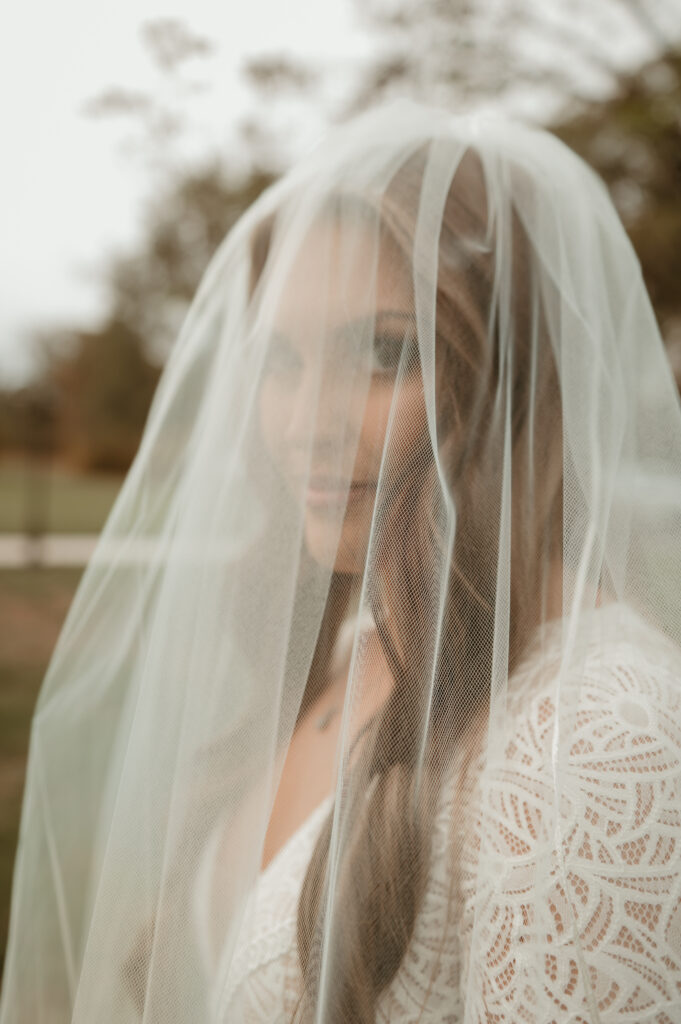An intimate portrait of a bride posing for our wedding photography services. The image captures the bride’s face partially obscured by a delicate, soft-focus tulle veil. She is wearing a white lace-detailed wedding dress, highlighting the intricate texture of her bridal attire. The composition focuses on her soft expression and the ethereal, romantic quality of the veil, providing a perfect example of our soft, cinematic photography style for wedding galleries.