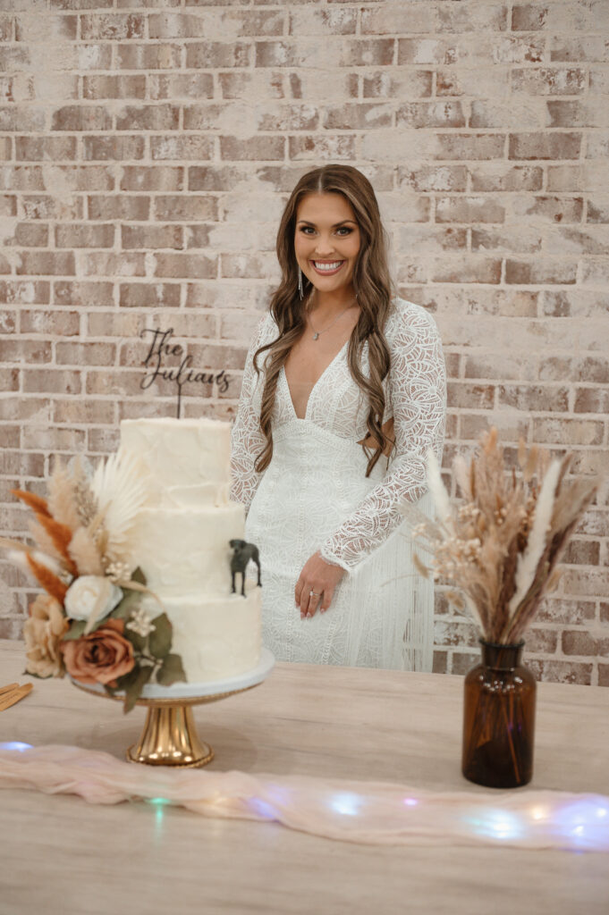 A smiling bride stands behind a three-tiered white wedding cake decorated with bohemian-style dried florals and a custom 'The Julians' cake topper, set against a rustic white brick wall. The cake features a unique dog figurine detail, showcasing personalized wedding cake design services.