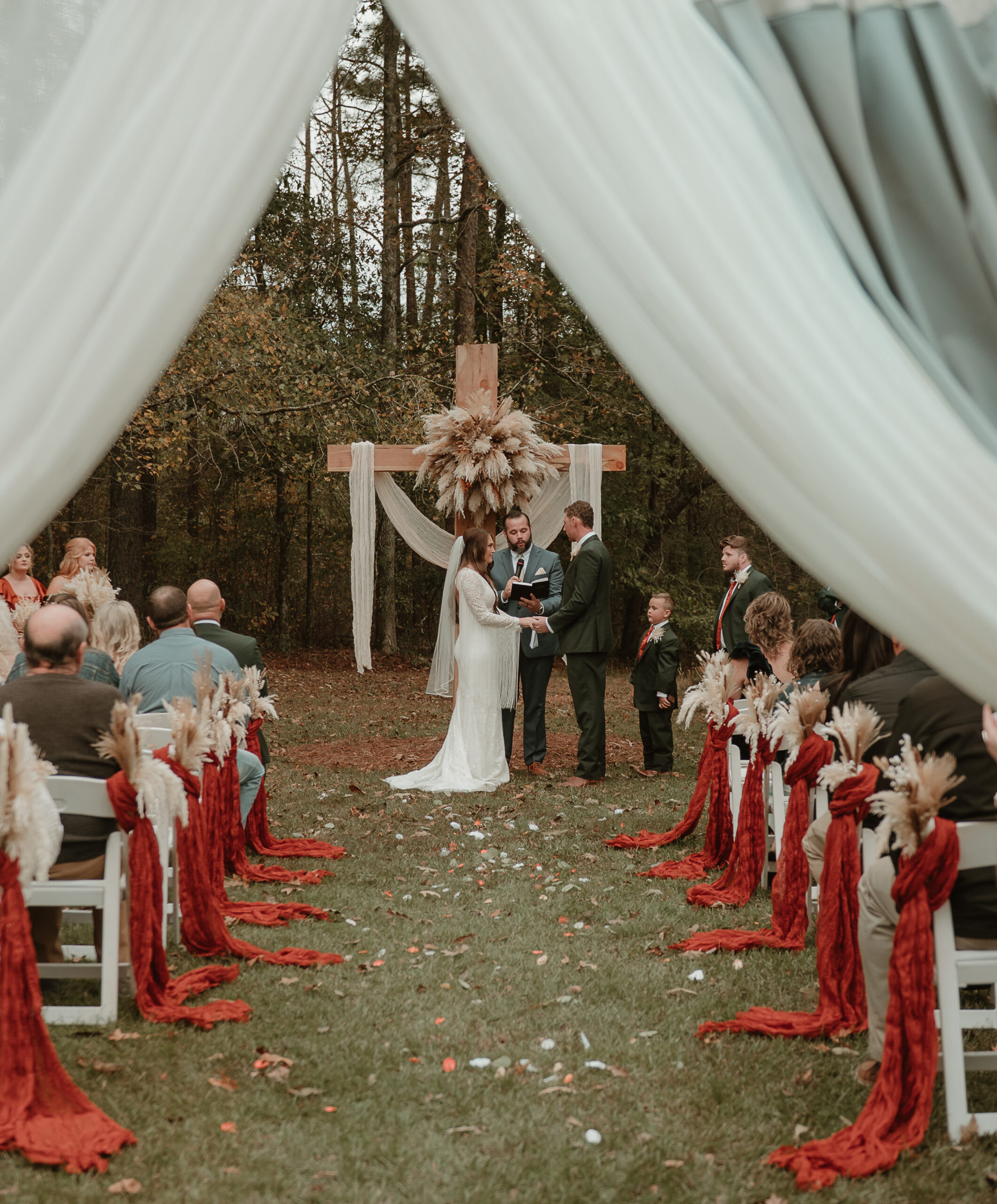 A framed view through white drapery captures an intimate outdoor wedding ceremony. The bride and groom stand before a wooden cross adorned with pampas grass and soft white linens, framed by tall autumn trees. Guests are seated on white chairs decorated with striking red fabric drapes and dried floral arrangements. This shot highlights our ability to curate romantic, rustic, and sophisticated ceremonies that blend natural forest beauty with thoughtful, thematic décor for a truly memorable exchange of vows.