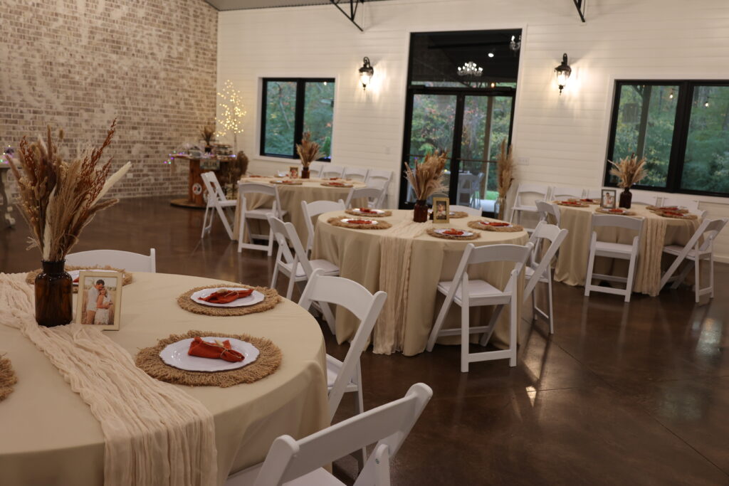 This image showcases our indoor venue set up for a warm, autumnal wedding reception. The round tables are draped in elegant, neutral-toned linens and feature matching table runners, rustic jute placemats, and burnt-orange napkins. Each centerpiece includes dried pampas grass arrangements and personalized photo frames, highlighting the intimate and inviting atmosphere we offer for special celebrations. The bright, white-walled space with its brick accent wall creates the perfect canvas for a sophisticated, cozy wedding design.