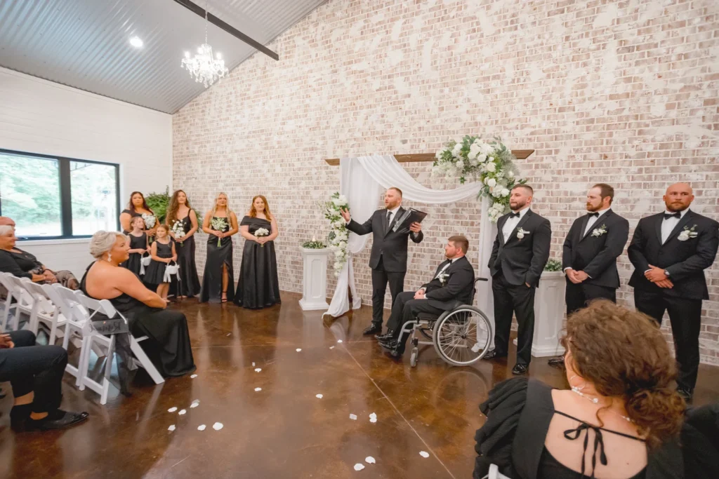 A wide-angle view of an indoor wedding ceremony in a rustic-chic event hall with a white-washed brick wall and polished concrete floors. A couple stands under a floral wooden arch with their wedding party, including a groom in a wheelchair.