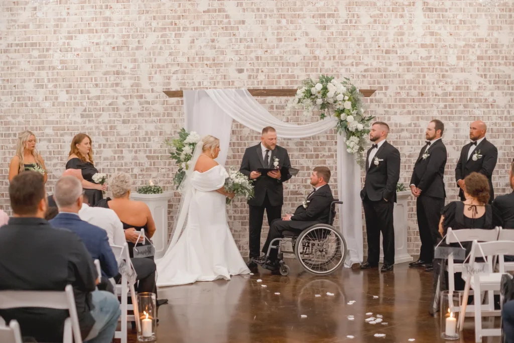 An intimate indoor wedding ceremony held in front of our signature whitewashed brick wall. The bride and groom stand at the altar with their officiant and wedding party, surrounded by white floral arrangements and elegant drapery. The rustic industrial aesthetic of the venue provides a warm, timeless backdrop for this couple’s vows, highlighting how our versatile space can be personalized to create a beautiful, accessible, and heartfelt ceremony for any couple.