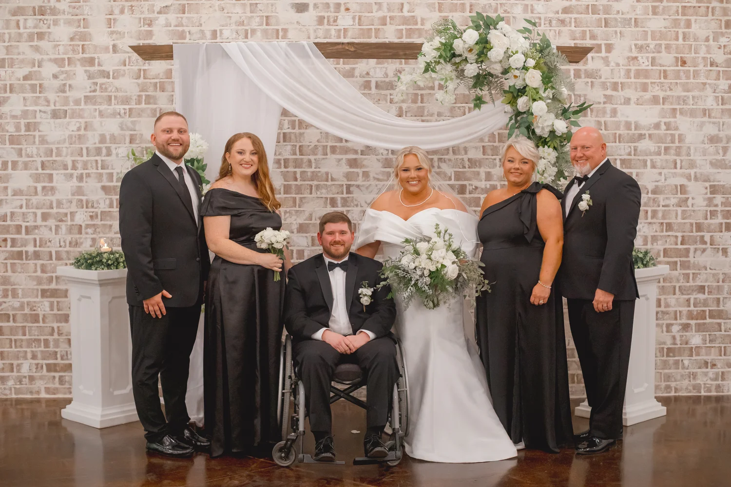 A formal wedding party portrait posed in front of a whitewashed brick ceremony backdrop with floral arrangements, showcasing the venue's modern aesthetic.