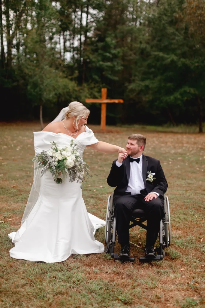 A tender, candid shot of a bride in a white gown and veil holding her groom’s hand as he sits in a wheelchair wearing a tuxedo. They are smiling at each other in an outdoor grassy field with a wooden cross in the background. This image perfectly captures the genuine love and connection between our clients during their wedding day ceremony.