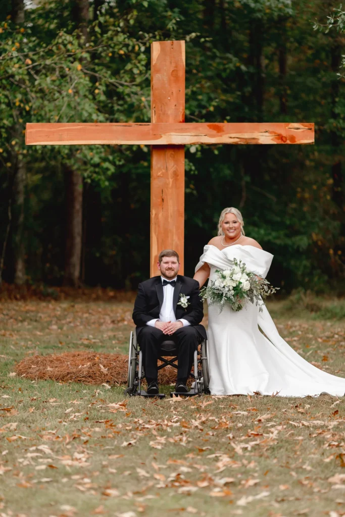 A bride and groom posing in front of a rustic wooden wedding cross at an outdoor forest venue, highlighting the inclusive and scenic atmosphere of Piney River Ranch.