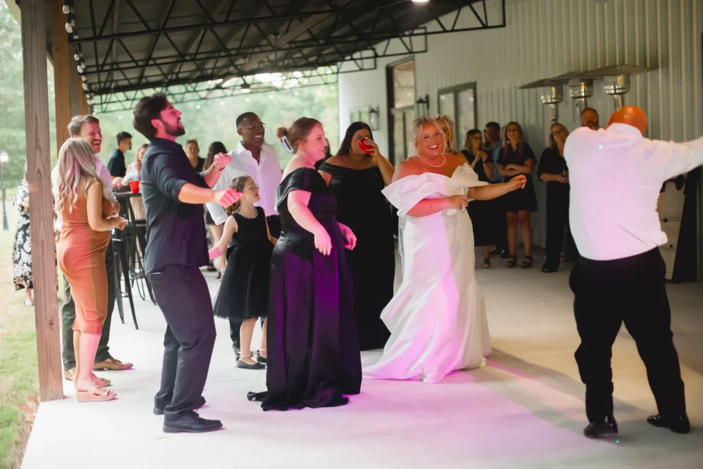 A joyful bride and wedding guests celebrate on our expansive covered patio at Lemon Ridge Events. This photo highlights the versatile outdoor space of our 10,000-square-foot Jasper facility, featuring industrial-chic architecture and warm string lighting. The scene captures the lively, effortless atmosphere our venue provides for larger celebrations of up to 250 guests. Whether for dancing or lawn games, this flexible area ensures a memorable reception for our Birmingham and Tuscaloosa area couples, reflecting our commitment to personal and seamless milestone events.