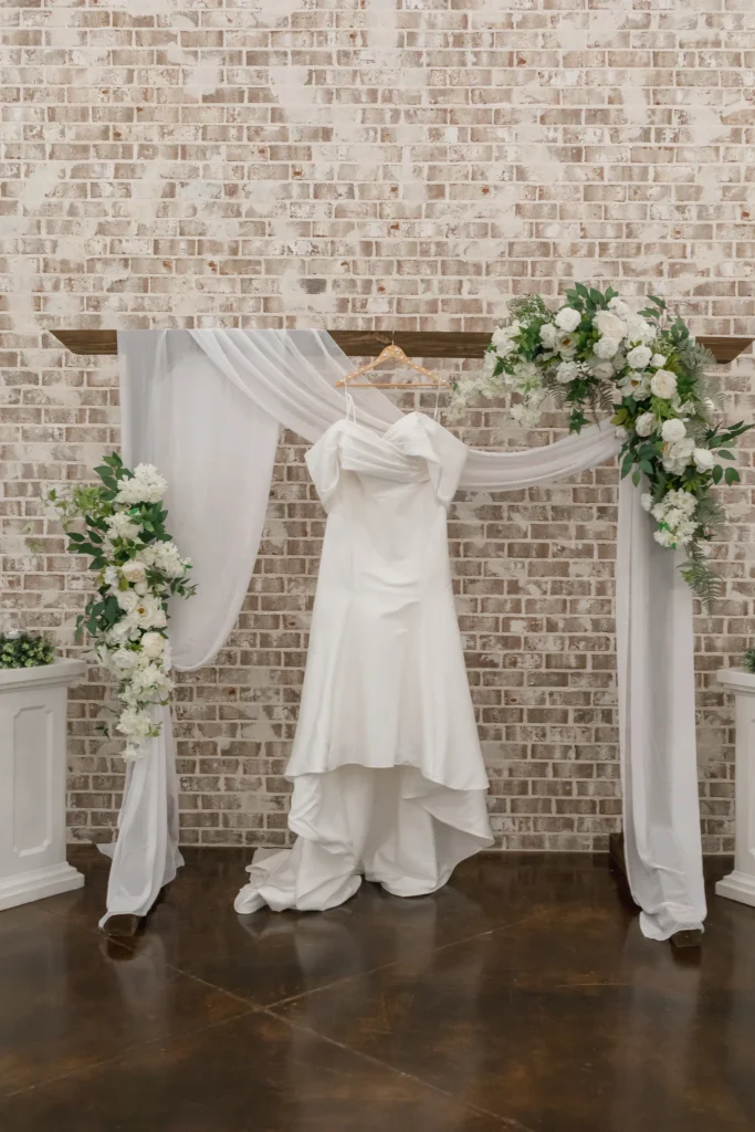 A classic white wedding dress hung on a wooden archway with floral arrangements and drapery in front of a rustic brick wall at a wedding venue.