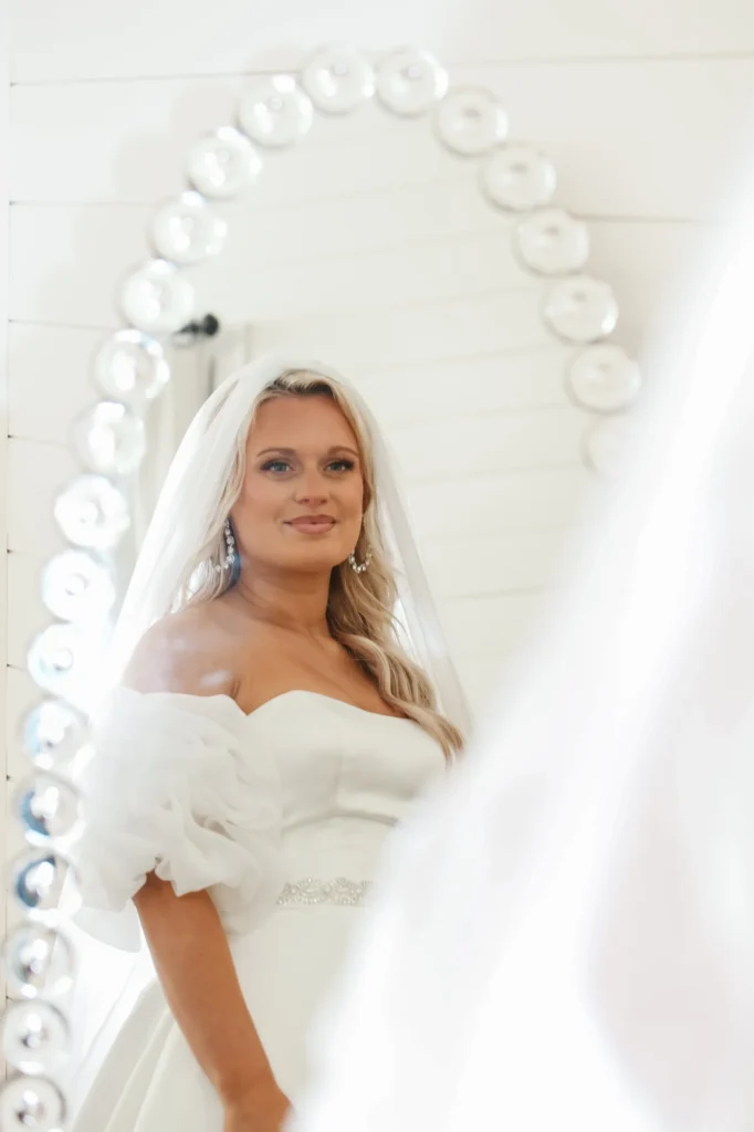 A portrait of a bride capturing her reflection in an oval, globe-lit mirror. The bride is wearing an off-the-shoulder gown with ruffled sleeves and a veil, looking directly into the glass. The soft, bright lighting emphasizes her bridal makeup and the elegant details of her dress. This image highlights our venue’s sophisticated preparation areas and the timeless, radiant moments we help our clients create on their wedding day.