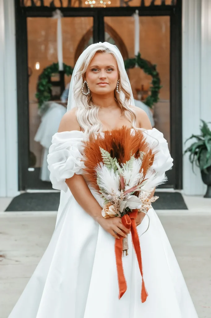 A portrait of a bride standing in front of glass doors wearing an off-the-shoulder wedding gown with a veil, holding a rustic bridal bouquet with pampas grass and burnt-orange foliage, showcasing an autumn wedding aesthetic.