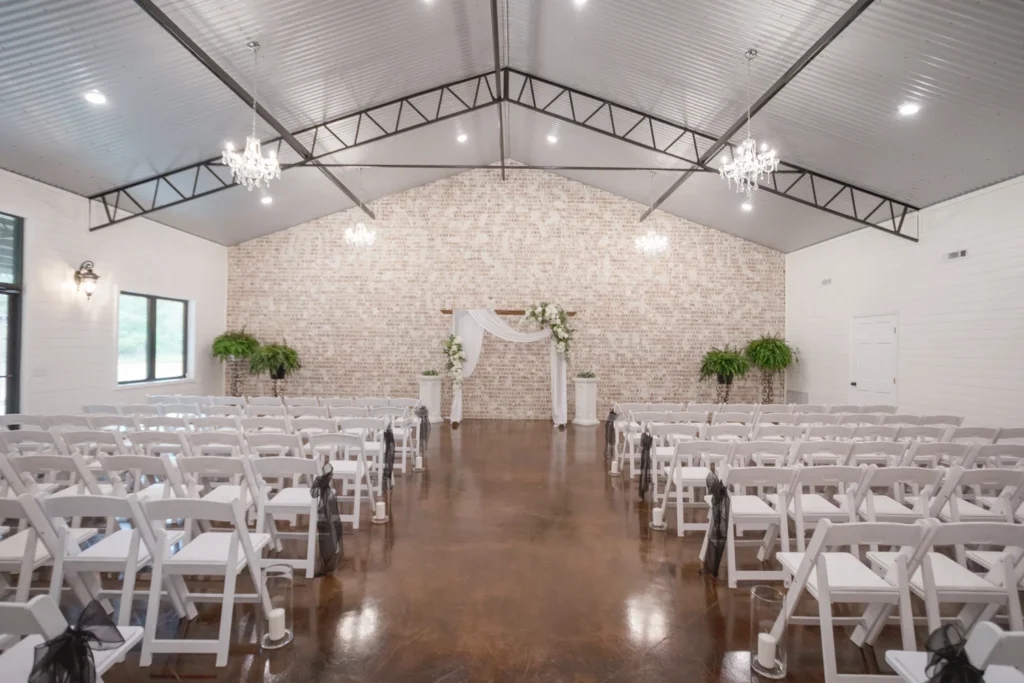 This image showcases our versatile indoor ceremony space at Lemon Ridge Events. The room features a stunning whitewashed brick focal wall, elegant chandeliers, and industrial-chic vaulted ceilings with exposed trusses. Arranged with our white folding chairs, the hall provides a sophisticated atmosphere for wedding ceremonies or formal gatherings. The polished floors and ample natural light highlight the room’s spaciousness, which can accommodate up to 250 guests. This climate-controlled facility ensures a beautiful, effortless setting for your milestone celebration, serving as a perfect year-round option for couples in the Jasper and Greater Birmingham areas
