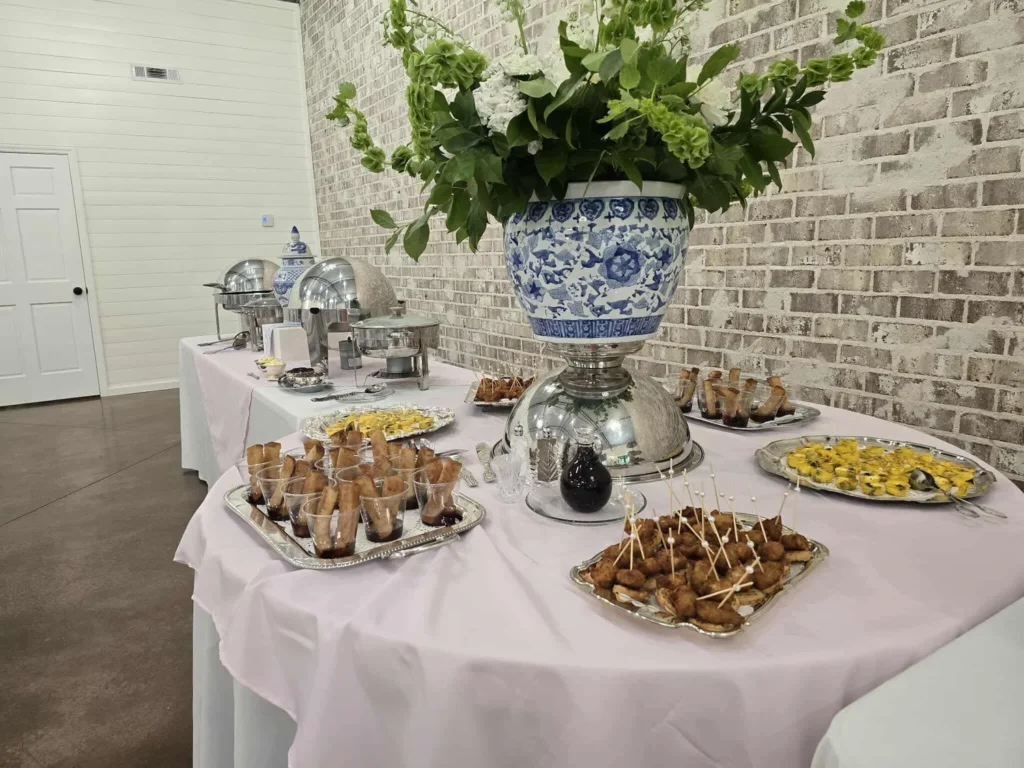 An elegant indoor reception buffet at Lemon Ridge Events featuring soft pink linens, silver serving platters, and a blue-and-white ginger jar centerpiece against white-washed brick walls.
