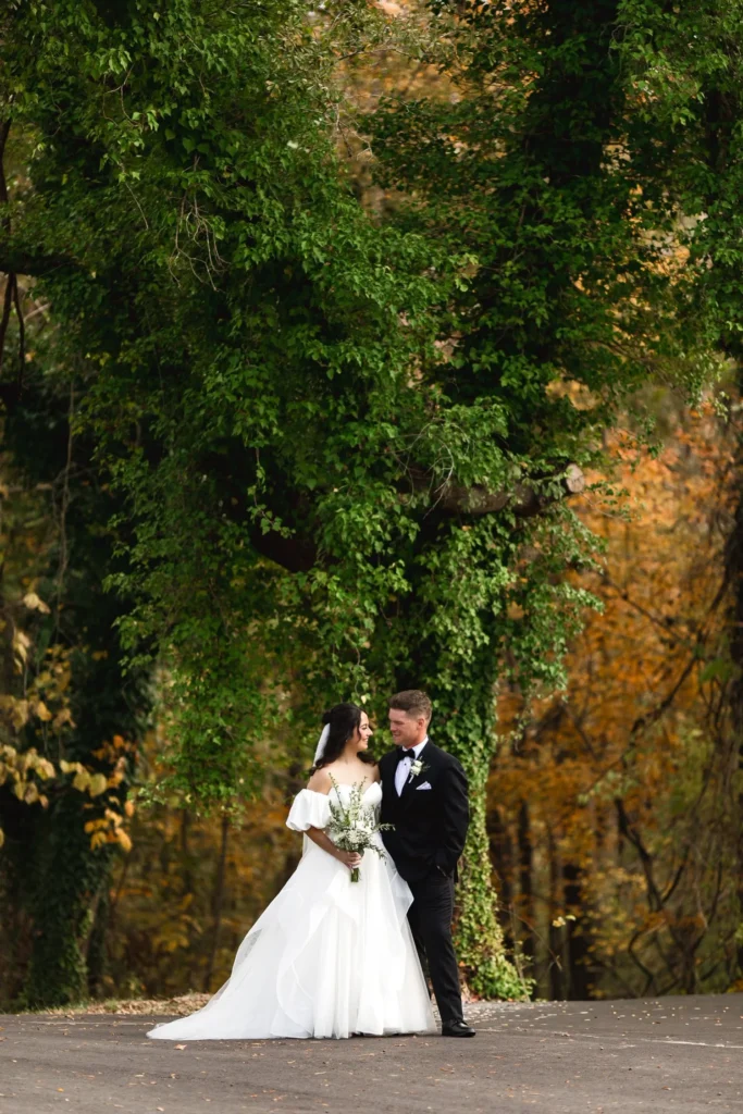 A vertical, full-body shot of a bride and groom standing on an asphalt path, facing each other lovingly. The bride wears an off-the-shoulder white wedding gown with a flowing skirt and holds a small bouquet. The groom wears a formal black tuxedo. They are framed by a large, lush tree draped in vibrant green ivy, set against a backdrop of golden autumn foliage. This image captures the romantic and natural aesthetic we prioritize for our fall wedding clients.