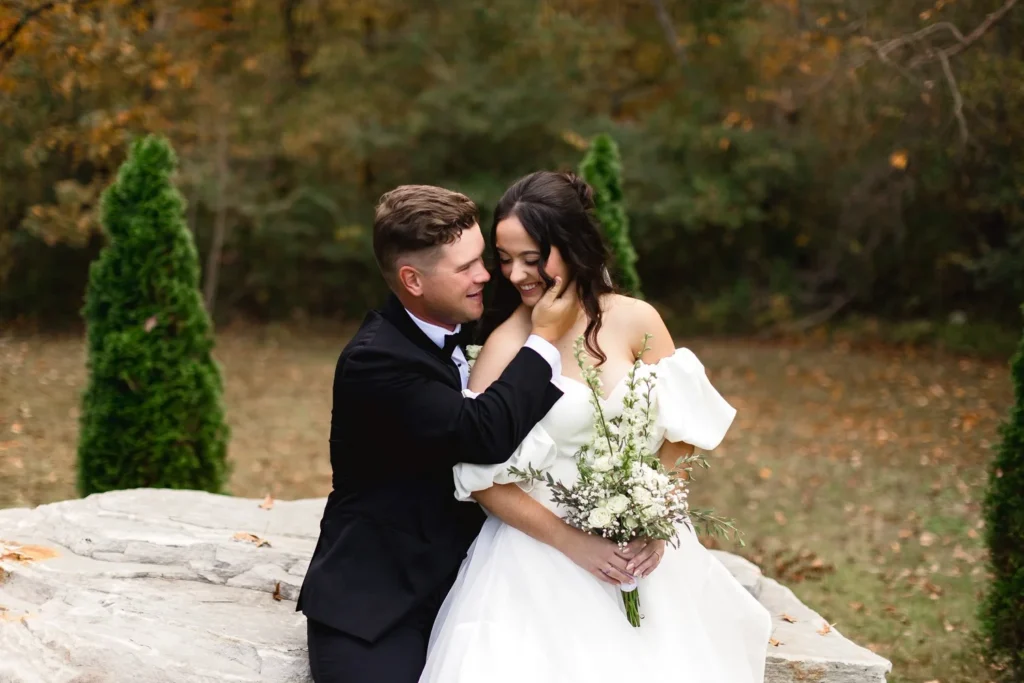A tender portrait of a bride and groom sitting on a stone ledge in an autumn forest, highlighting the wedding venue's scenic outdoor photo opportunities. The groom wears a black tuxedo and the bride wears an elegant white off-the-shoulder gown while holding a white bouquet.