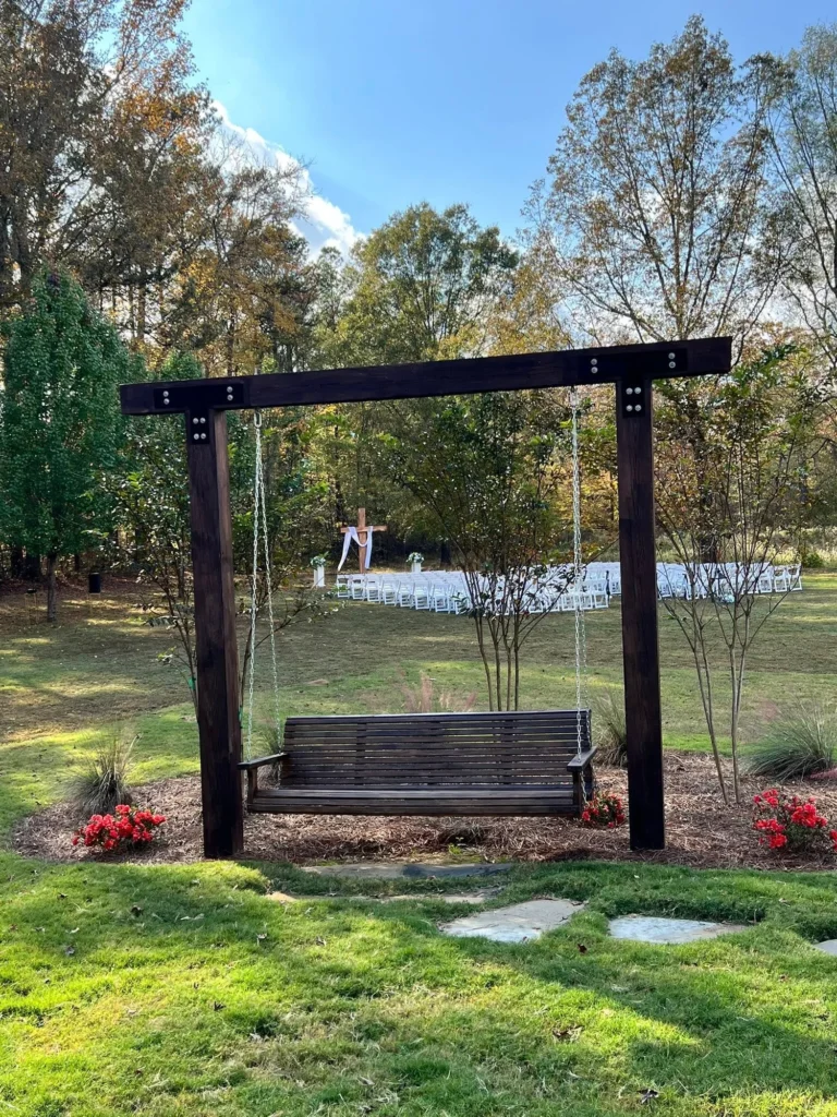 A view from our grounds showing a large wooden garden swing in the foreground, serving as an inviting vantage point. In the background, an outdoor wedding ceremony site is set up with white chairs facing a wooden cross altar, nestled among mature trees. This image perfectly captures the serene, natural beauty of our outdoor wedding space and the peaceful atmosphere we offer for couples exchanging vows.