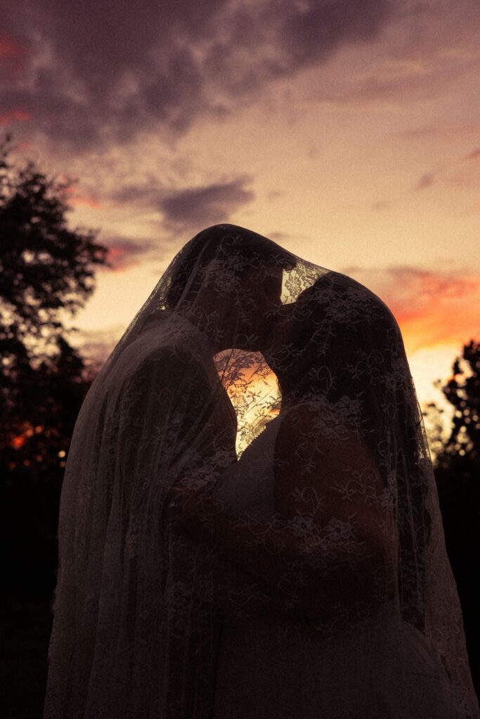 A romantic silhouette of a bride and groom sharing a kiss beneath a lace bridal veil at sunset, highlighting the intimate and artistic quality of wedding photography.