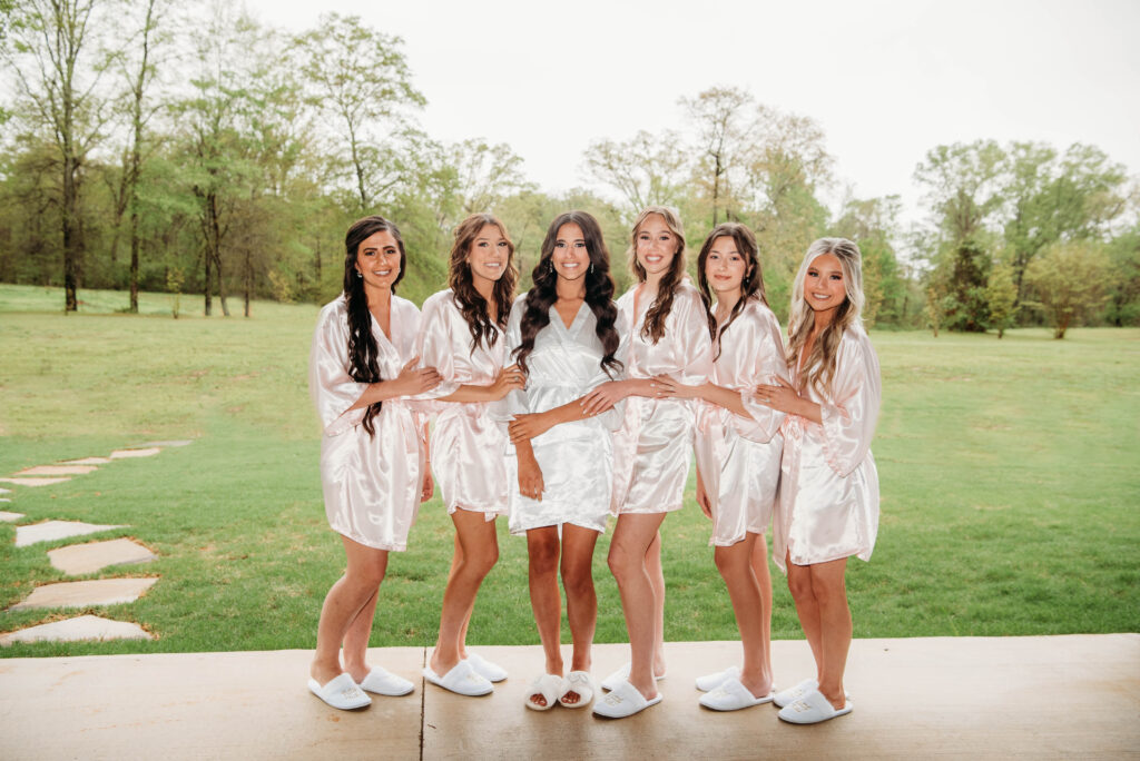This image features a radiant bride in a white robe standing center stage with her five bridesmaids, who are dressed in coordinating soft pink silk robes and matching slippers. They are positioned on a patio overlooking our lush, green outdoor ceremony grounds. This shot perfectly captures the intimate, relaxed morning energy of wedding day preparations before the main event begins.
