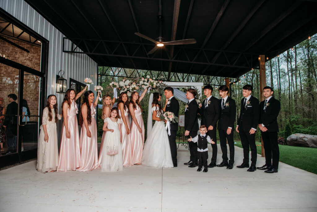 A full wedding party poses under a covered outdoor patio area. The bride and groom stand center, surrounded by bridesmaids in blush pink gowns and groomsmen in black suits, with a flower girl and ring bearer in the foreground. The group is positioned in front of a signature wedding arch with a lush floral arrangement. The image captures the spacious, modern-industrial aesthetic of the outdoor ceremony space during golden hour.