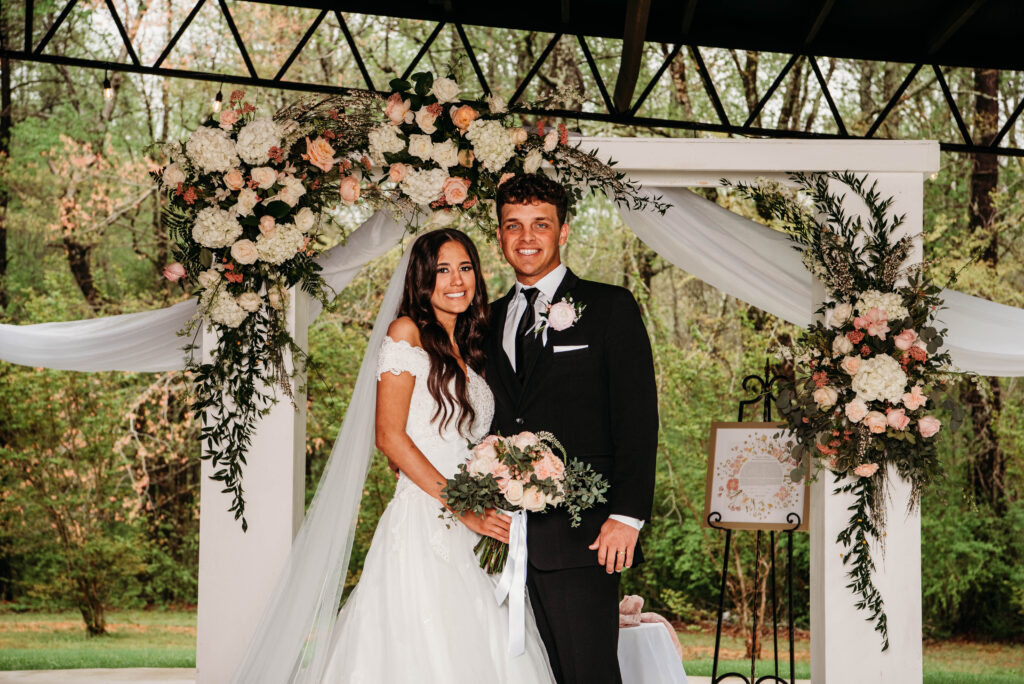 A portrait of a newlywed couple smiling under a custom-decorated white floral wedding arch at a scenic outdoor woodland venue.