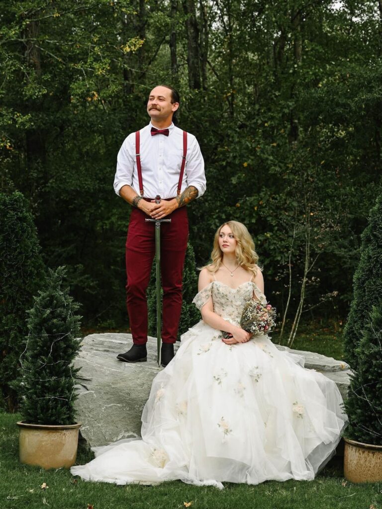 A bride and groom pose in a whimsical, enchanted forest clearing at Willow Creek Wedding Venue. The groom stands on a mossy boulder holding a sword, while the bride sits in a floral-detailed gown, highlighting the venue's storybook-themed outdoor wedding offerings.