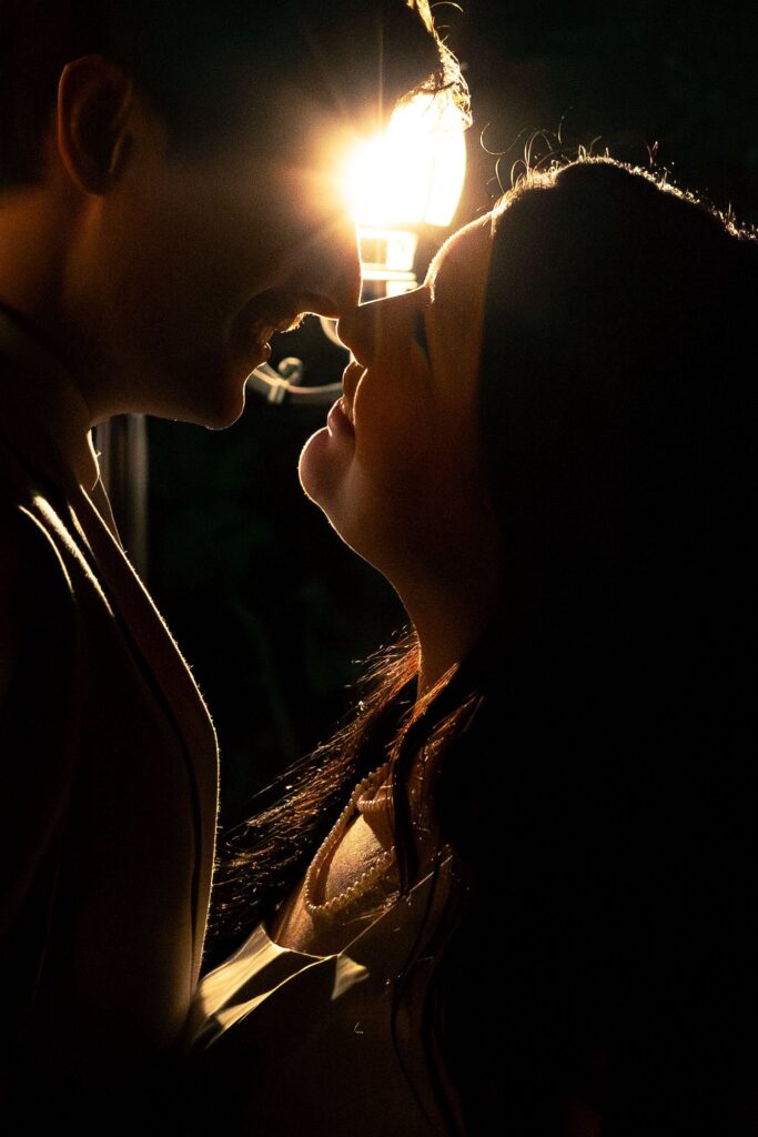 A intimate, backlit portrait of a couple sharing a tender moment at night. The light source creates a soft, golden rim light around their profiles, emphasizing their closeness. This image captures the romantic, atmospheric essence of our wedding photography services, perfect for showcasing how we immortalize the quiet, emotional highlights of a couple's special day.