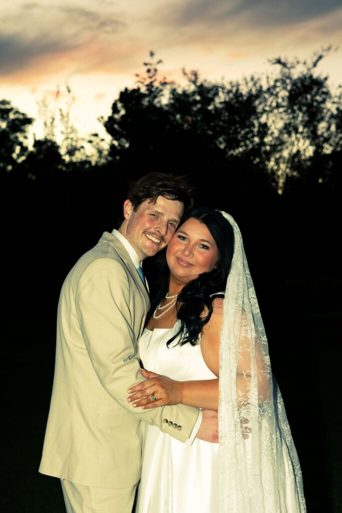 A close-up portrait of a bride and groom embracing during golden hour. The bride wears a classic white gown with a delicate lace veil, and the groom is dressed in a light-colored suit. The background features a vibrant sunset sky silhouettes against trees, capturing an intimate, romantic moment from one of our wedding events. This image perfectly highlights the emotion and joy we strive to facilitate for every couple at our venue.