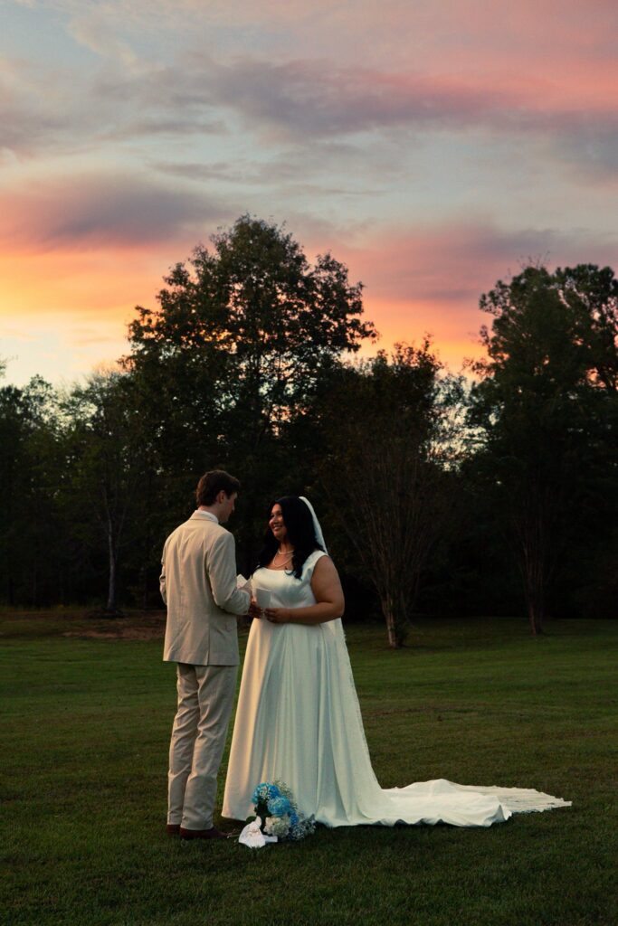 A bride and groom stand facing each other in an open, grassy field during the golden hour. The bride wears a long white wedding dress with a veil, and the groom is dressed in a light-colored suit. They are holding small notes, captured in a quiet moment of sharing vows. A small floral bouquet rests on the grass near the bride’s train. The background features lush trees silhouetted against a vibrant, colorful sunset sky, highlighting the romantic and intimate atmosphere of their wedding day.