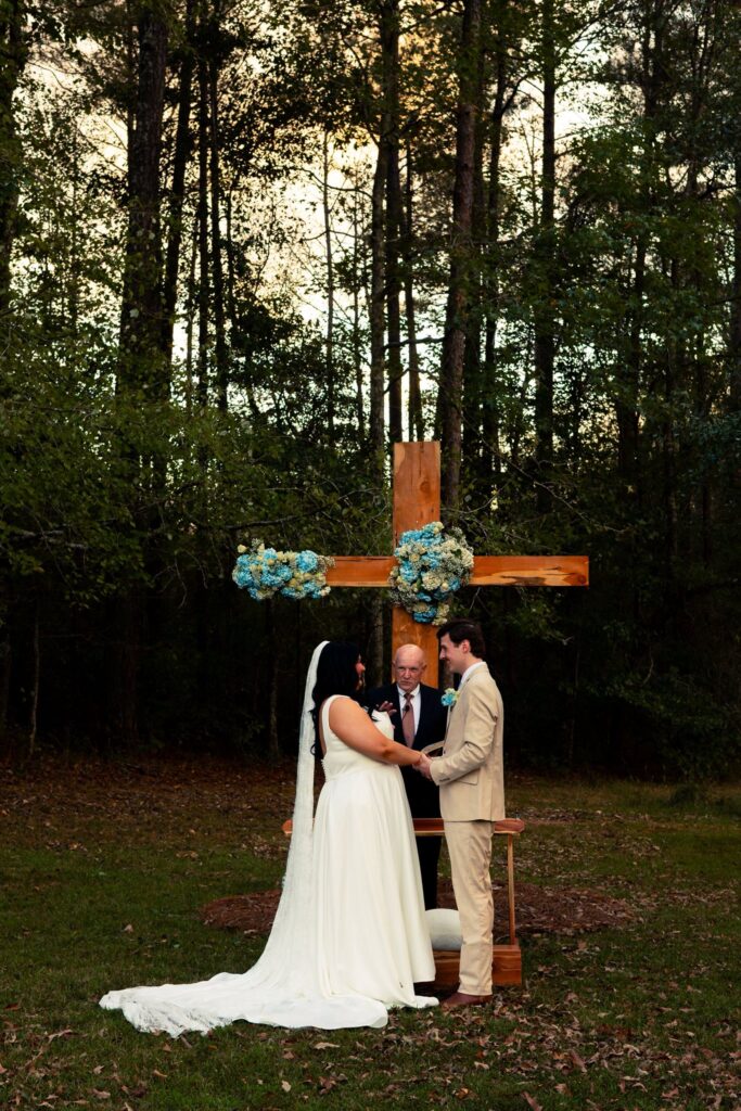 A bride and groom holding hands during their wedding ceremony at our outdoor venue. They are standing in front of a rustic wooden cross adorned with blue and white floral arrangements, with a forest backdrop. An officiant stands between the couple, presiding over their vows in an intimate, wooded setting.