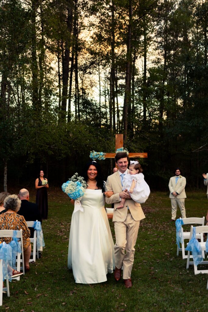 A joyful bride and groom walking down the aisle after their outdoor ceremony at Evergreen Woods, with the groom carrying their infant daughter. They are surrounded by family and friends seated in white chairs, set against a backdrop of dense forest trees and a wooden cross altar. The scene captures a quintessential, intimate woodland wedding at our venue, highlighting the natural beauty of the forest setting during the golden hour.