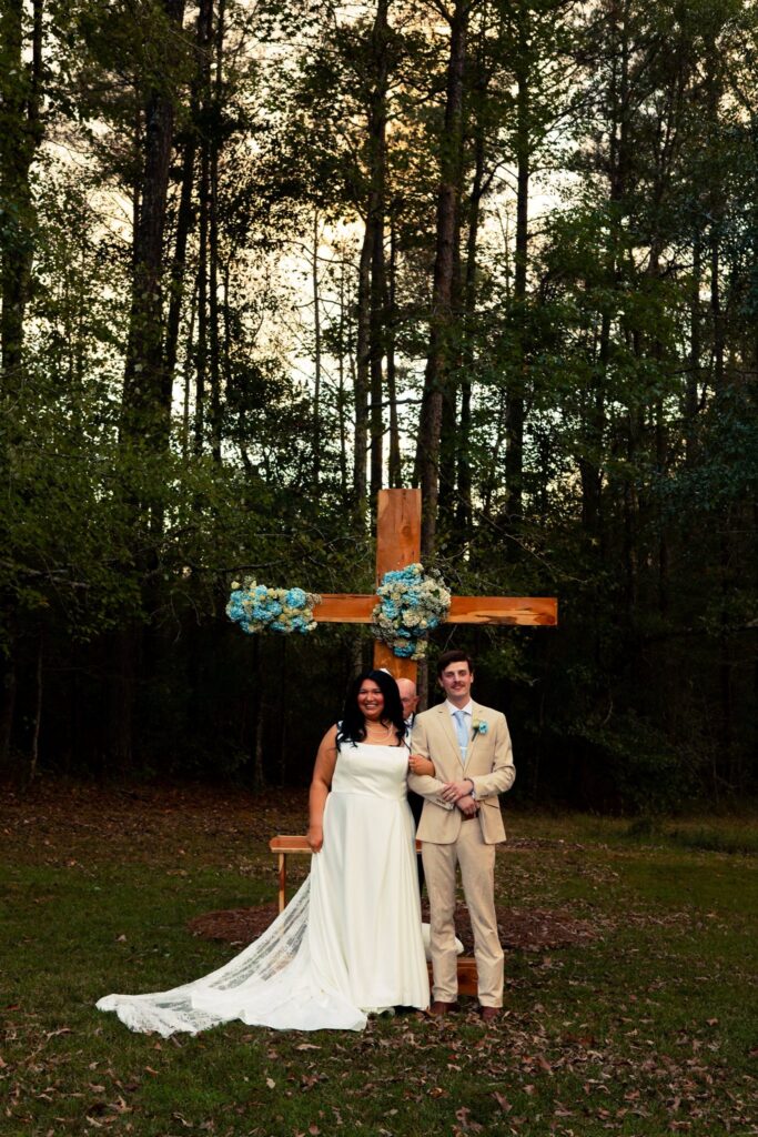 A newly married couple standing in front of a custom-built wooden cross adorned with floral arrangements. The ceremony takes place in a tranquil, wooded outdoor setting, highlighting the natural beauty and intimate atmosphere our venue provides for faith-centered weddings.