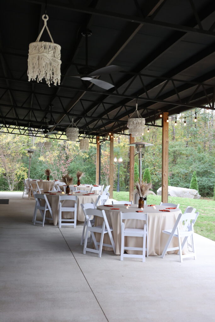 This image features our covered outdoor pavilion arranged for a wedding reception. Round tables are dressed in cream linens with dried floral centerpieces and white folding chairs. The industrial-style ceiling is decorated with bohemian macramé pendant lights and string lights, providing a warm atmosphere. The space opens up to our natural, wooded landscape, offering a perfect blend of modern comfort and rustic charm for guests to enjoy throughout the event.