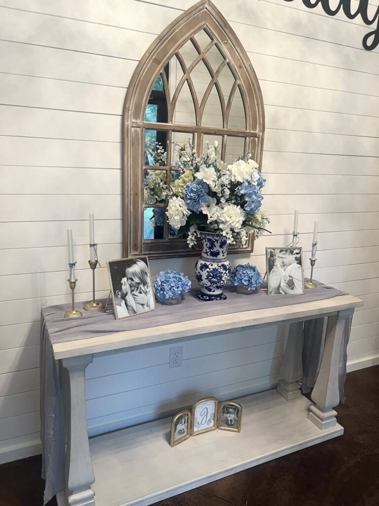A rustic console table setup featuring a blue and white hydrangea floral arrangement in a decorative vase, flanked by framed wedding photos and elegant taper candles. The arrangement is centered under an arched wooden cathedral mirror against a white shiplap wall, creating a sophisticated focal point for guests. A draped blue runner and small framed signage on the lower shelf complete this welcoming display, perfect for showcasing personalized wedding details or memorial photos at our venue.