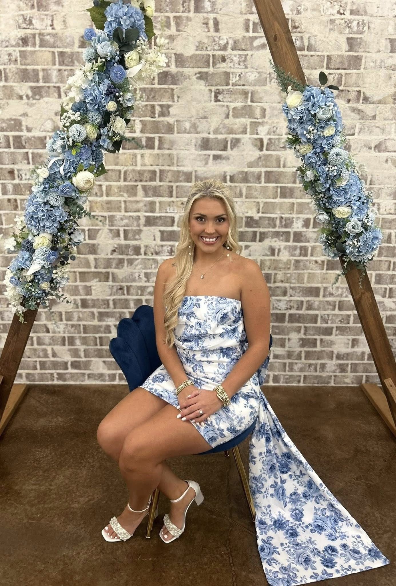 A client posing in front of our signature wooden triangular arch, adorned with elegant blue and white floral arrangements. The setting features a rustic brick backdrop, perfectly complementing the client's blue-and-white floral dress and matching accessories. This image showcases our event space's versatility for sophisticated bridal showers, engagement photos, or special occasion portrait sessions.