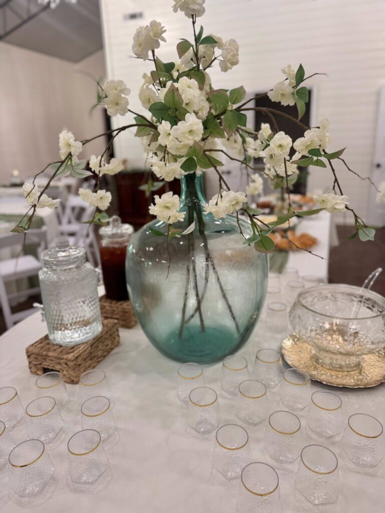 A beautifully styled beverage station centerpiece featuring white cherry blossom branches in a large, teal-tinted glass vase. The table is set for guests with an array of gold-rimmed glasses, glass dispensers, and a crystal punch bowl. This elegant setup adds a touch of organic charm to our event hosting space, perfect for spring-themed weddings or bridal showers. It reflects our commitment to sophisticated, detail-oriented decor that enhances the guest experience.