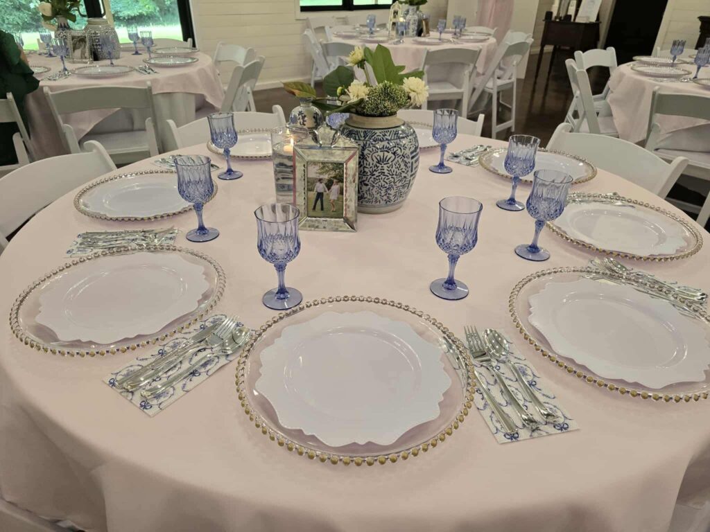 A meticulously curated round table setup for a wedding reception, featuring a soft pink tablecloth accented by blue-and-white chinoiserie vases and elegant blue glassware. The place settings include ornate beaded chargers, polished silverware, and custom patterned napkins, centered around a personal framed photo. This arrangement showcases our attention to detail and ability to execute sophisticated, theme-driven event decor in our event space.