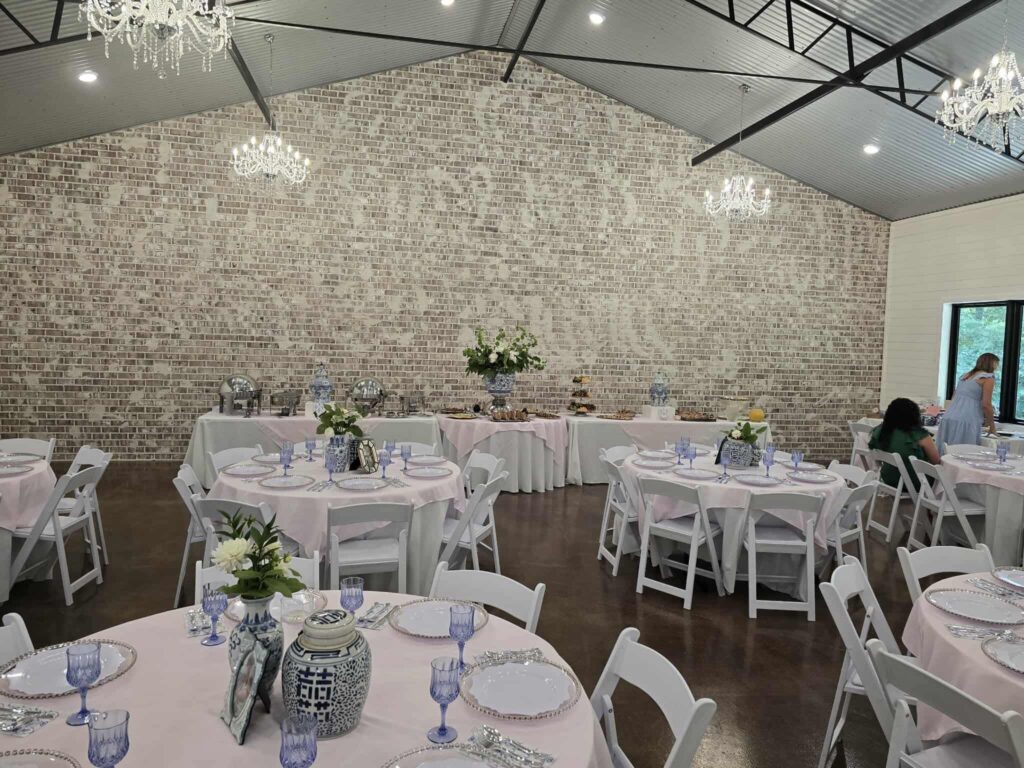 A wide-angle view of our main ballroom fully prepared for a wedding reception. The room features our signature whitewashed brick wall, crystal chandeliers, and multiple round guest tables draped in soft pink linens. Blue and white porcelain ginger jars serve as floral centerpieces, complemented by matching blue glassware. In the background, long buffet tables are set with catering service. This setup highlights our venue’s capacity for elegant, intimate dining experiences and our ability to accommodate sophisticated, themed decor.