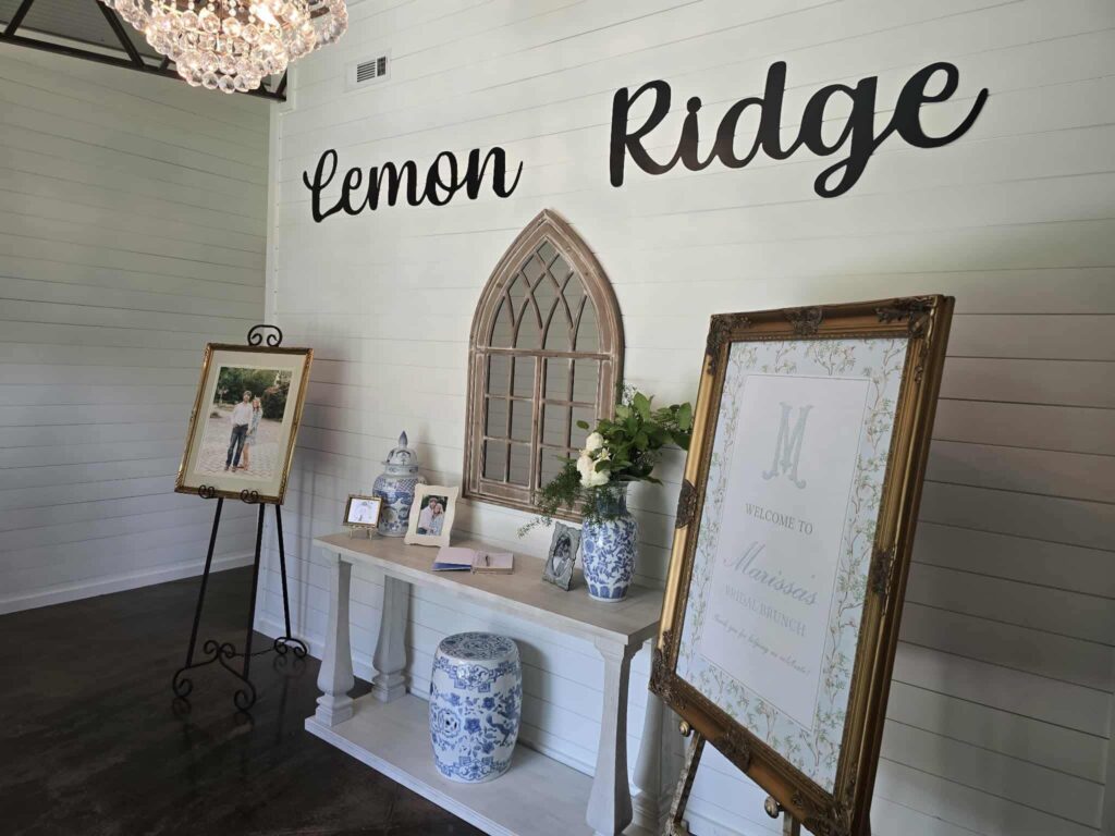The main entryway gallery wall at Lemon Ridge Wedding and Event Venue, featuring a personalized bridal brunch welcome sign, a couple's portrait on an easel, and a decorated console table with blue-and-white porcelain and floral arrangements.