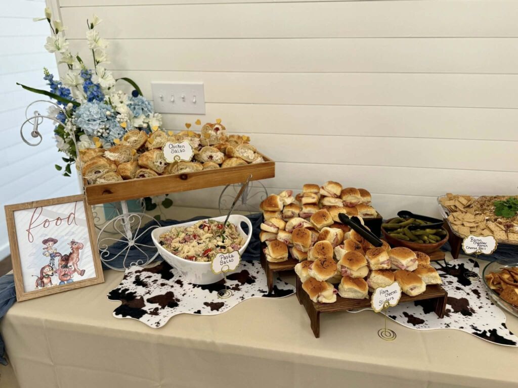 This catered food station features a rustic, western-themed display perfectly suited for themed events. The table includes a variety of finger foods, including chicken salad croissants, ham and cheese sliders, pasta salad, and a bacon ranch cheese ball, all presented on wooden risers with cow-print accents. A decorative "Toy Story" themed sign adds a playful touch to the professional catering setup, demonstrating our ability to coordinate menu presentation with specific event themes.