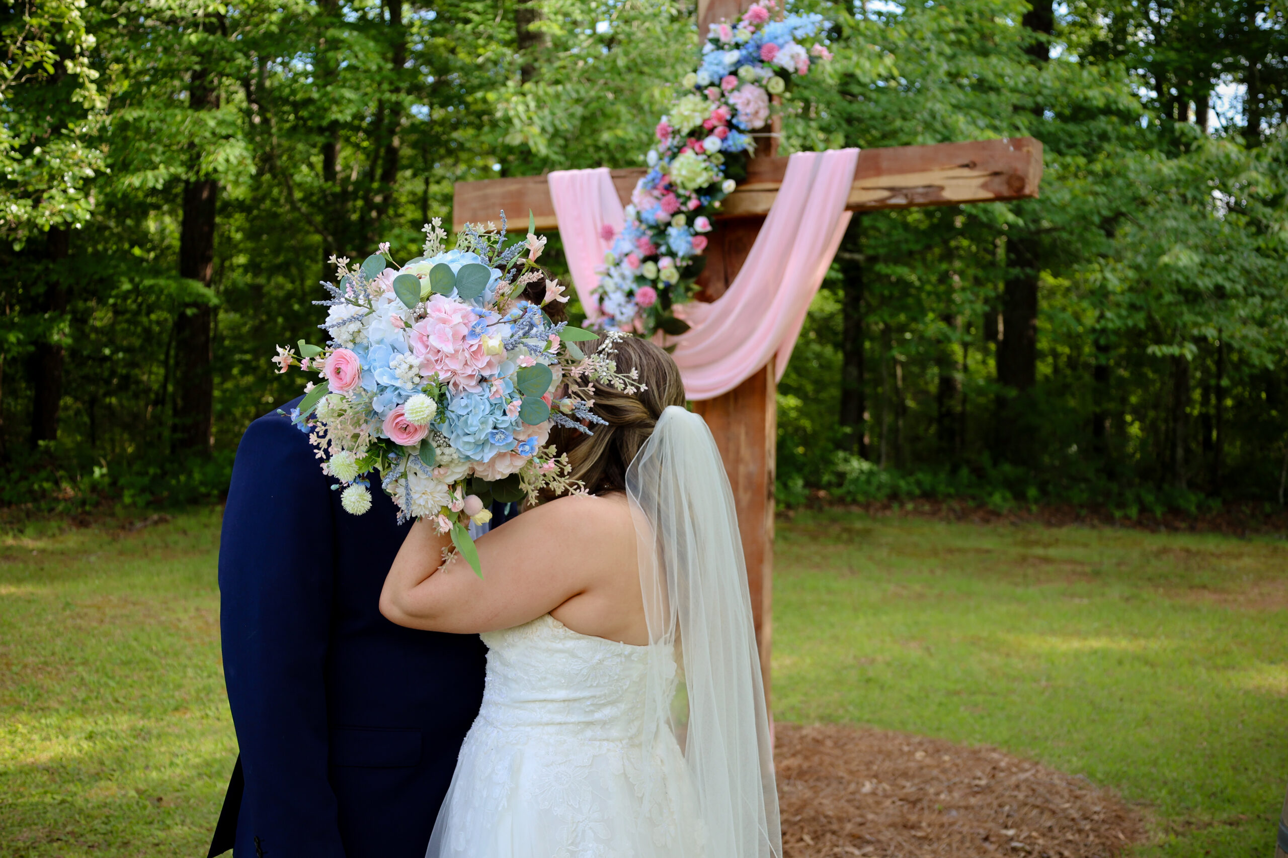 A romantic shot of a bride and groom sharing a private moment behind a large bridal bouquet. They are standing directly in front of our signature wooden cross altar, which is elegantly adorned with pastel floral arrangements and soft pink drapery. The lush green forest background highlights the peaceful, intimate atmosphere of our venue, perfectly framing the couple during their outdoor wedding ceremony.