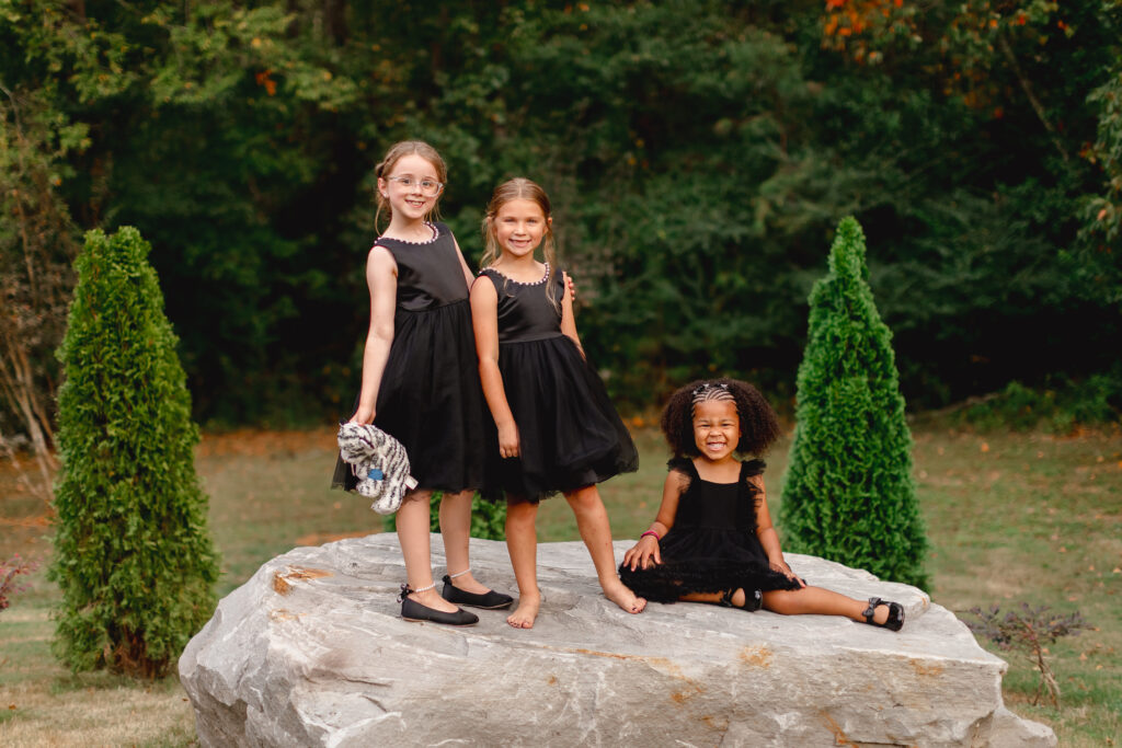 Three young guests in formal black attire pose on a large natural rock feature at one of our lush outdoor ceremony locations. This scene highlights the beautiful, versatile photography backdrops available on our Lemon Ridge Events property in Jasper, Alabama. Our expansive grounds provide the perfect setting for capturing candid family moments and wedding party portraits against a backdrop of mature trees and greenery. Whether hosting a micro wedding or a full celebration, our venue offers effortless opportunities to document every joyful milestone of your event.