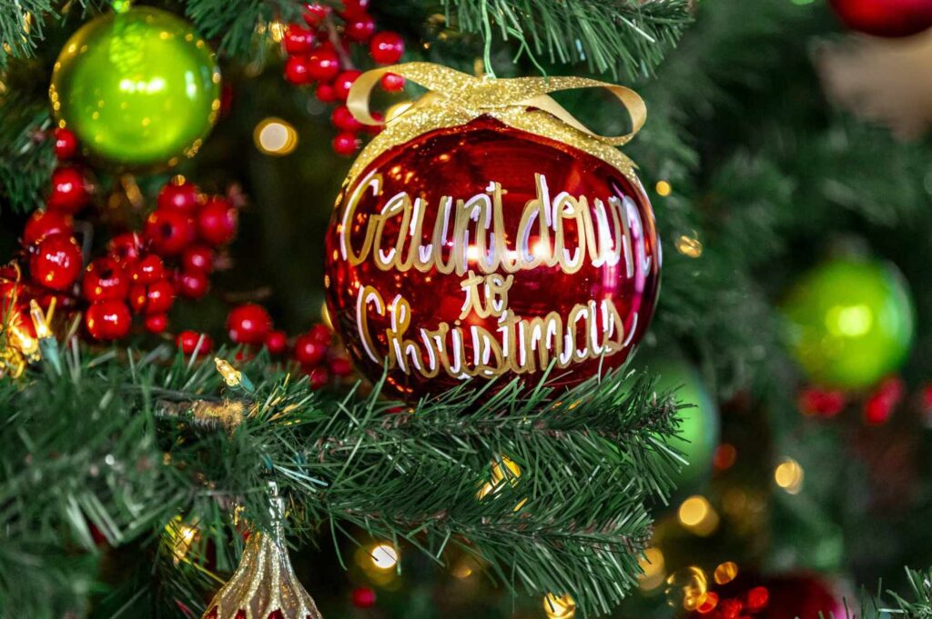 A close-up of a vibrant red ornament featuring "Countdown to Christmas" in gold script, nestled within a decorated evergreen tree. Surrounded by lime green baubles and clusters of red berries, this festive detail represents the warm, celebratory atmosphere at Lemon Ridge Events during the winter season. Our Jasper venue offers a beautifully decorated setting for holiday parties, winter weddings, and seasonal showers, ensuring a personal and effortless backdrop for every milestone celebration in the Birmingham and Tuscaloosa areas.