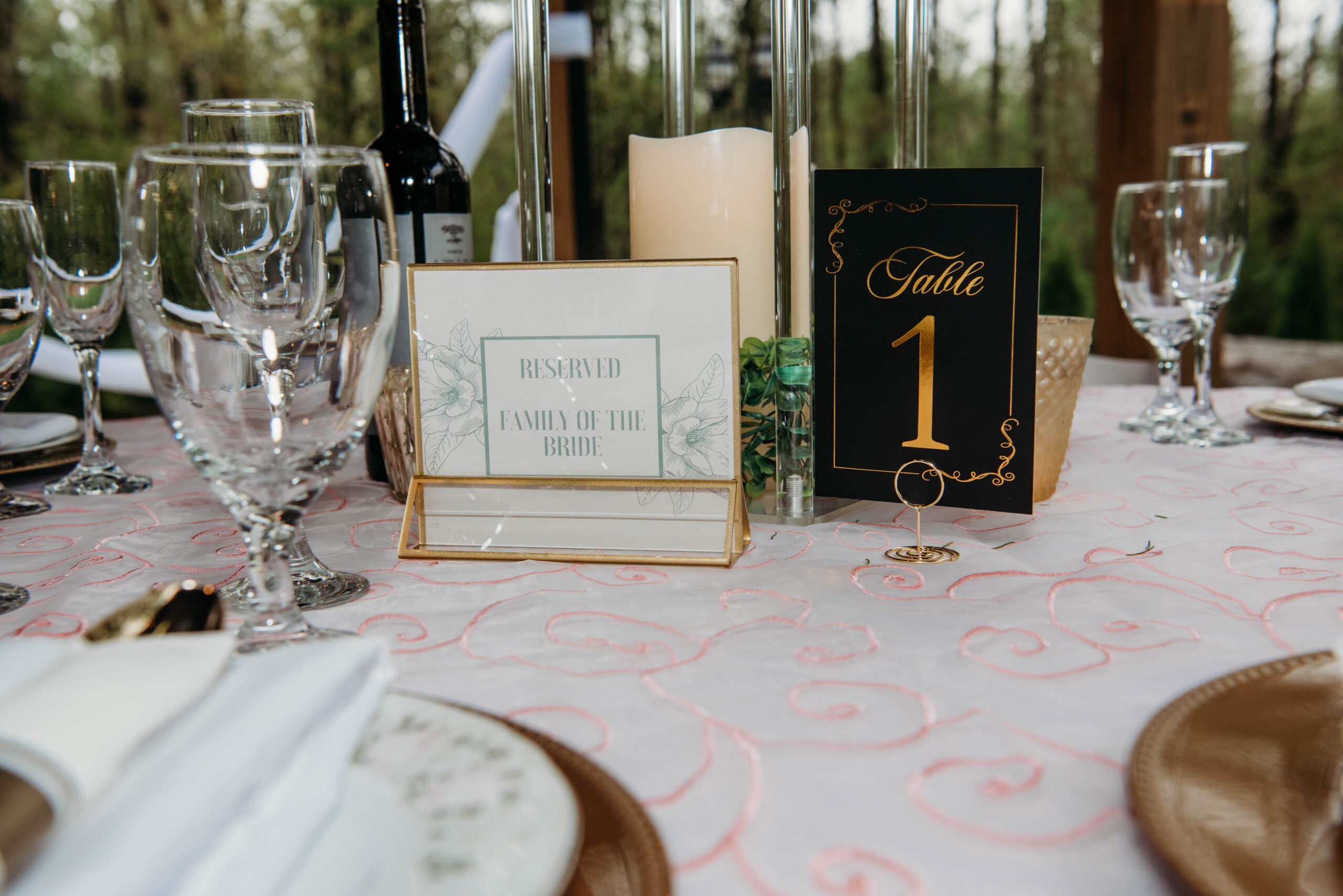 A close-up of a sophisticated reception table setting at Lemon Ridge Events. The scene features a white linen with pink swirl embroidery, elegant glassware, and a bottle of wine. Centrally displayed are a gold-framed “Reserved Family of the Bride” sign and a sleek black and gold “Table 1” card. This image highlights the personalized attention to detail and effortless organization we provide for our couples in Jasper. Our versatile venue accommodates these refined touches, ensuring every VIP guest feels welcomed and every seating arrangement is perfectly executed.