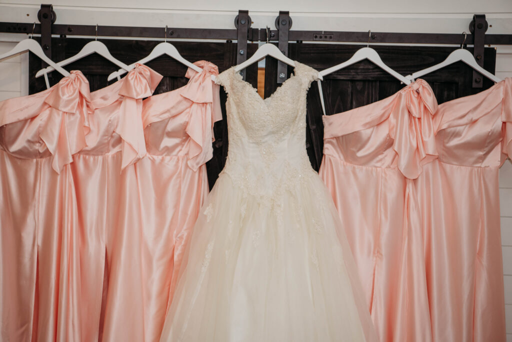 This image showcases a stunning lace wedding gown flanked by four shimmering peach bridesmaid dresses, hanging from a rustic barn door track against clean white shiplap. Taken within one of our dedicated bridal suites at Lemon Ridge Events, this scene highlights the elegant, photo-ready spaces we provide for wedding preparations. Our 10,000+ square foot facility in Jasper offers these intentional details, ensuring that every moment—from getting ready to the final send-off—is both effortless and beautiful for our couples and their wedding parties.