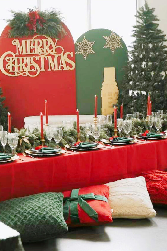 This elegant holiday-themed display showcases the versatility of our 10,000+ square foot facility for seasonal celebrations. The setup features a long table with a vibrant red cloth, evergreen garland, red taper candles, and sophisticated place settings with green napkins and gold chargers. A custom "Merry Christmas" backdrop with red and green arched panels, festive greenery, and decorative floor pillows creates an inviting atmosphere. This demonstrates how Lemon Ridge Events can be transformed into a festive, immersive space for holiday parties, winter weddings, or seasonal reunions in the Jasper area.