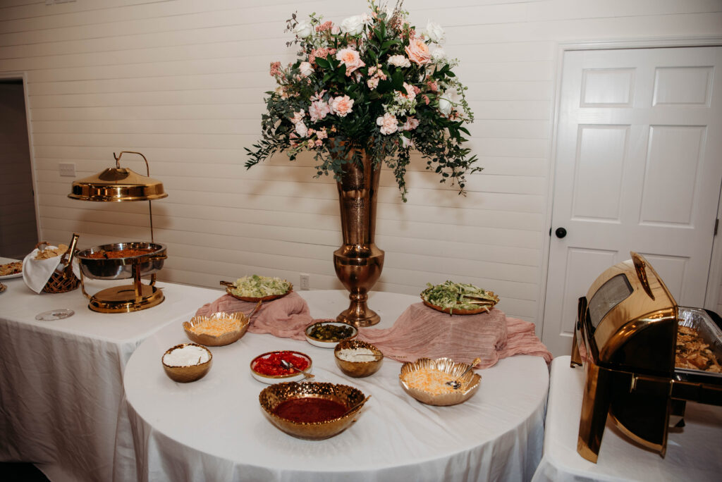 This image showcases a professionally styled catering station at Lemon Ridge Events, featuring polished gold chafing dishes and hammered gold serving bowls. A tall, ornate gold vase with a lush pink and white floral arrangement serves as a sophisticated centerpiece against our signature white shiplap walls. This setup highlights the versatility of our 10,000+ square foot facility, which easily accommodates various dining styles for weddings and parties. With full kitchen access provided, your chosen caterer can provide seamless service, ensuring every detail of your milestone event is both beautiful and effortless.
