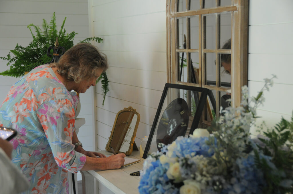A guest signs a commemorative book at a personalized welcome station inside Lemon Ridge Events. Positioned against our clean shiplap walls, the display features custom decor, including a framed vinyl record and blue floral arrangements. This image highlights the versatile indoor space of our 10,000-square-foot Jasper facility, which allows for unique personal touches at weddings, reunions, and parties. We offer a flexible canvas for every celebration, ensuring guests feel supported and welcomed from the moment they arrive at our Greater Birmingham area venue.