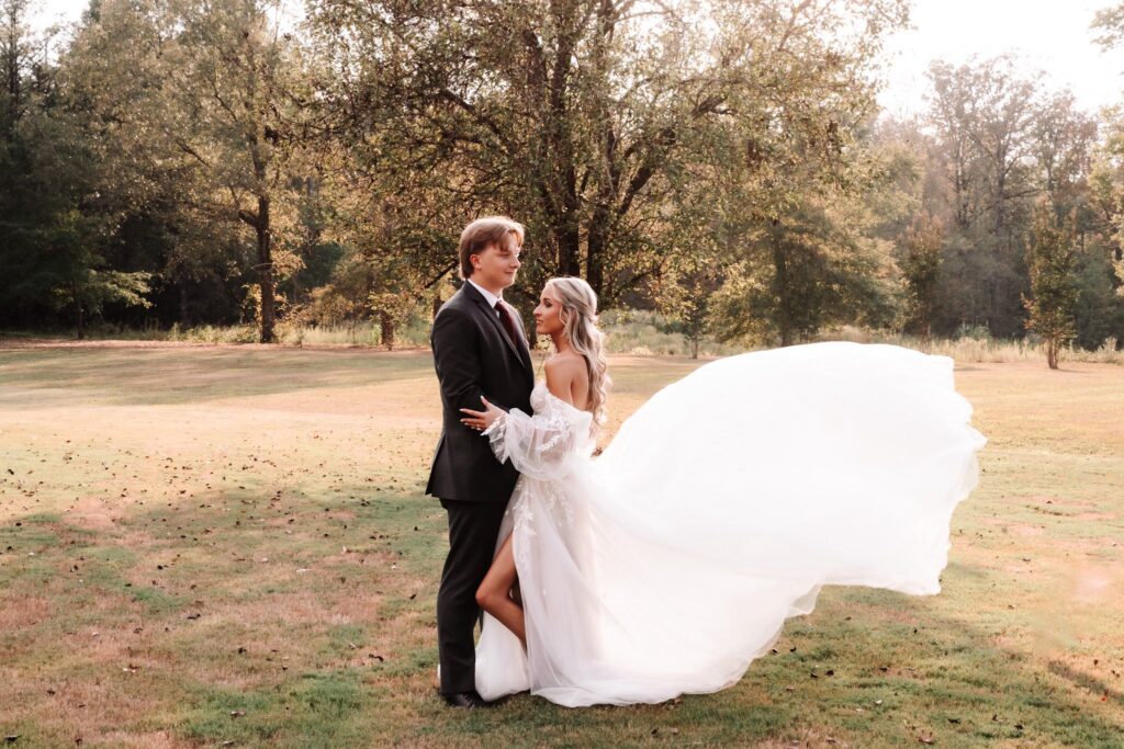 A stunning outdoor wedding portrait captured at Lemon Ridge Events, highlighting our versatile natural grounds. The couple stands in a sun-drenched field against a backdrop of lush trees, showcasing the romantic atmosphere our Jasper venue provides. The bride’s gown, featuring a dramatic flowing train, catches the soft breeze, illustrating the elegance and effortless beauty of our outdoor ceremony locations. This scene perfectly represents the personalized, memorable celebrations we host for couples in the Greater Birmingham and Tuscaloosa areas seeking a beautiful, flexible space for their milestone moments.